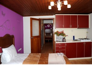 Ein Schlafzimmer mit einem hölzernen Kopfteil, einem Bett mit weißer Bettwäsche und einem Nachttisch mit einer roten Lampe. Ein Flur führt zu einem Badezimmer mit violetter Wand und einer Duschkabine. Die Küche verfügt über rote Schränke, einen Gasherd und eine Spüle mit einem Wasserkocher auf der Arbeitsfläche.