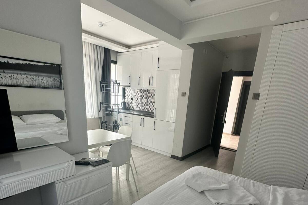 A modern hotel room with a large bed, a desk, a chair, and a kitchenette in the background.