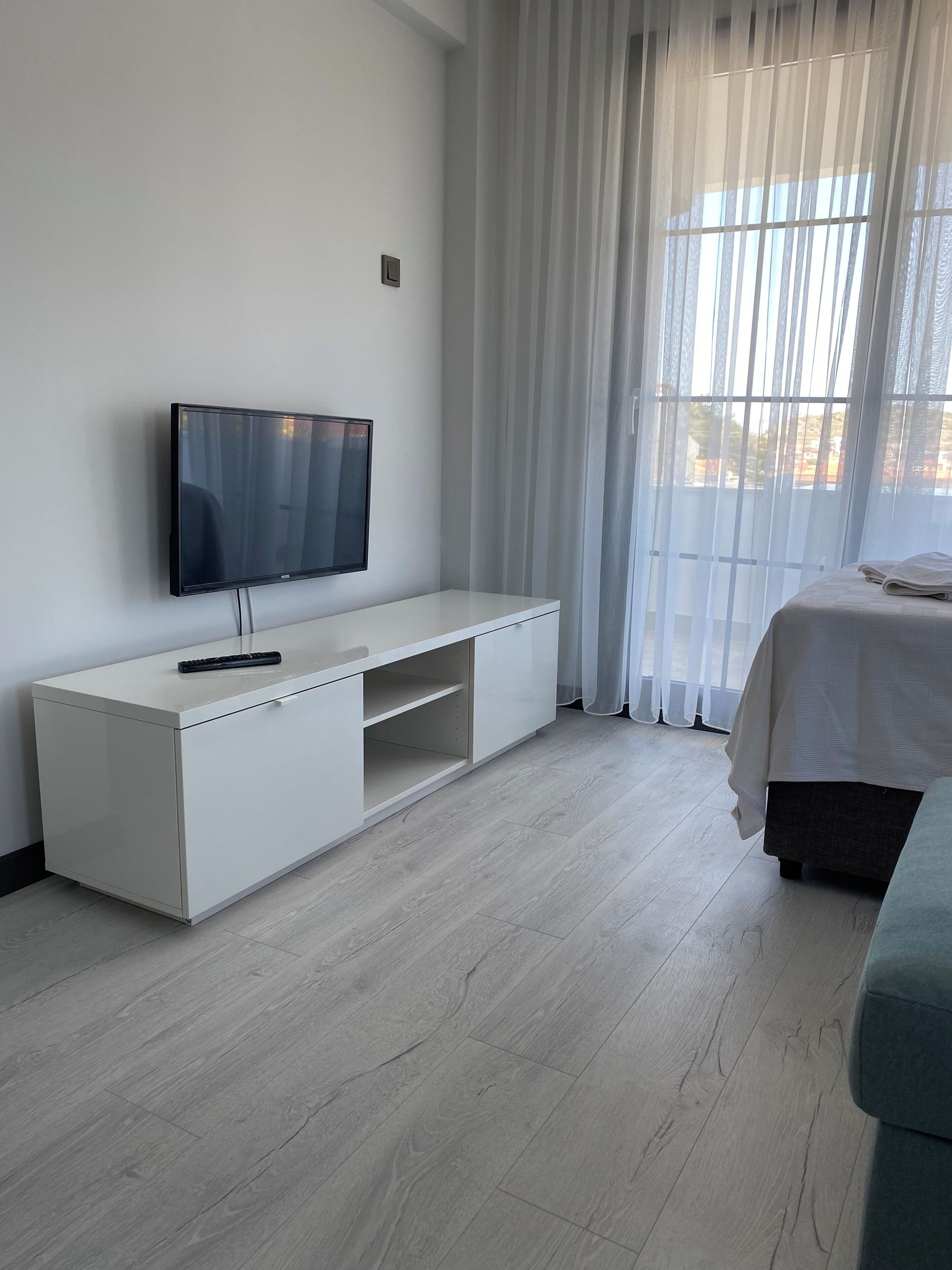 A modern hotel room with a flat-screen TV mounted on a white cabinet, a bed with a white duvet, and a large window with sheer curtains.