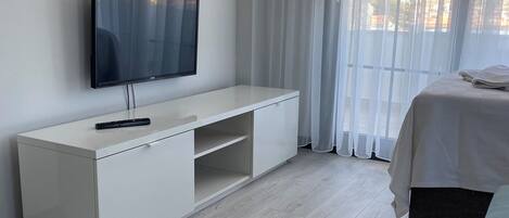A modern hotel room with a flat-screen TV mounted on a white cabinet, a bed with a white duvet, and a large window with sheer curtains.