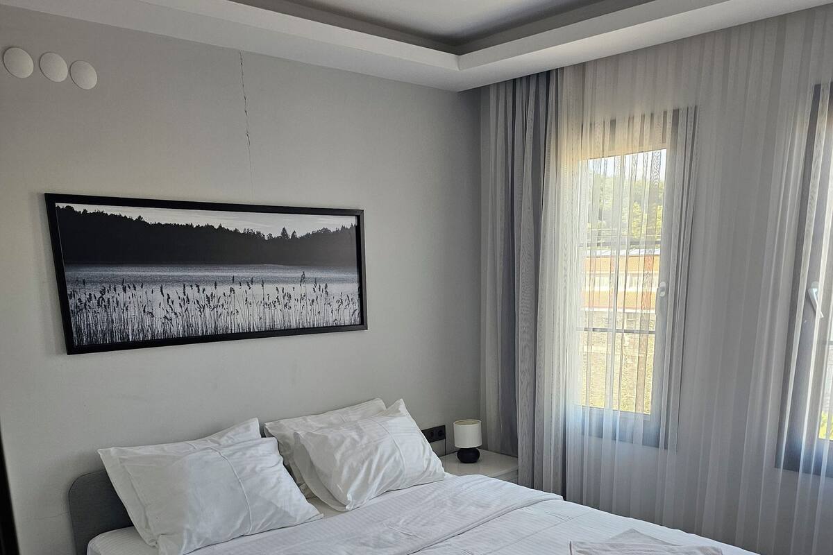 A neatly made bed with white linens, a large framed picture above it, and a window with sheer curtains.