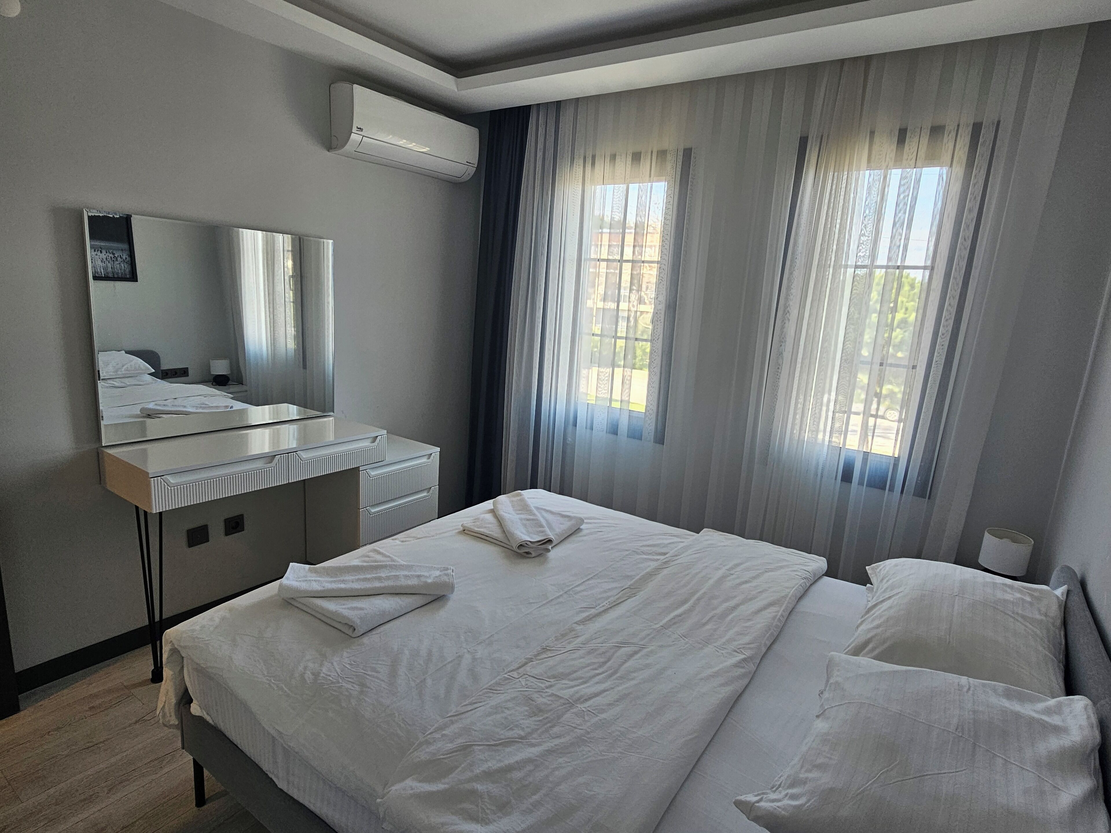 A hotel room with a bed, a bedside table, a mirror, an air conditioning unit, and a window with curtains.