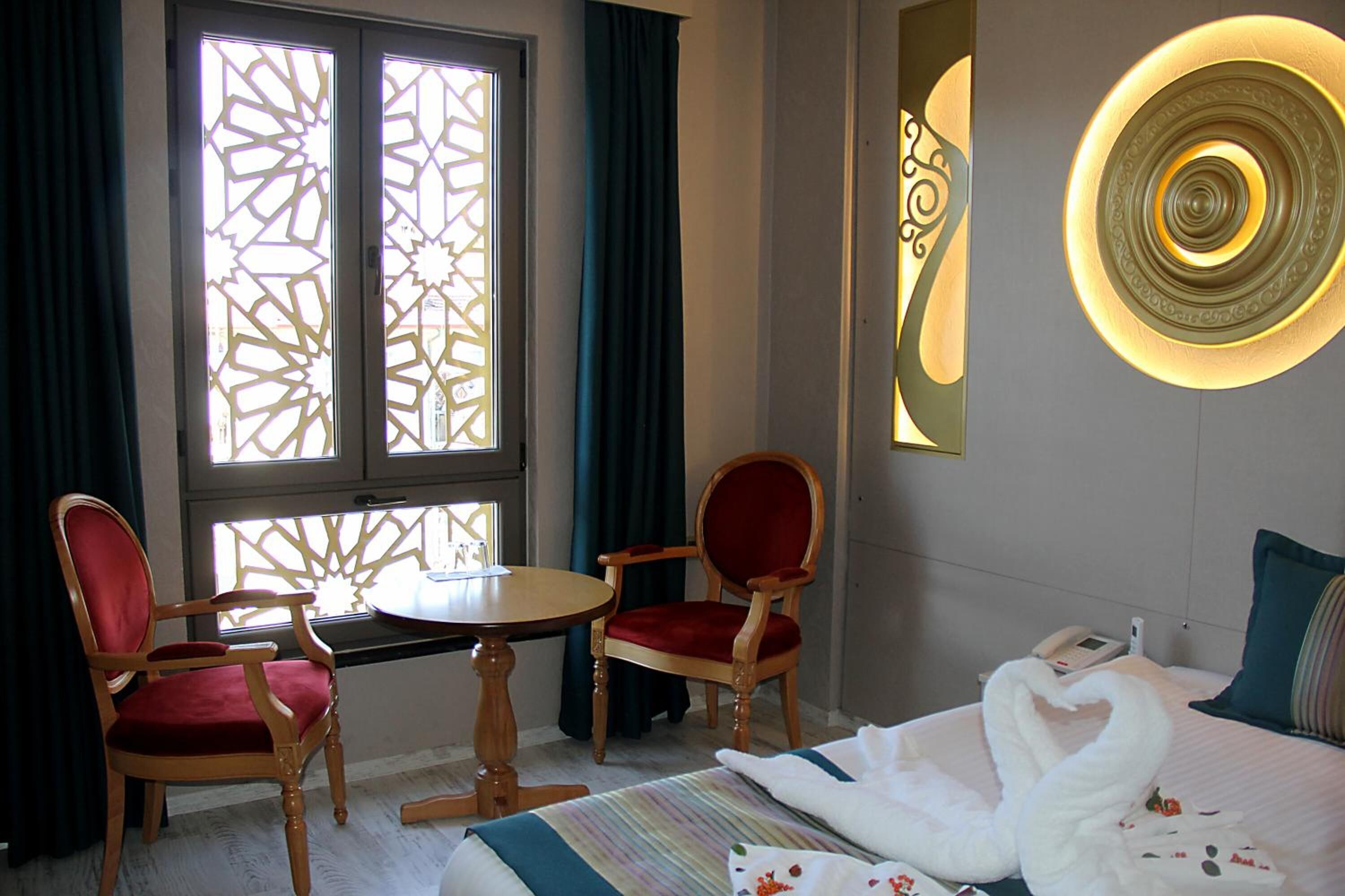 Habitación de hotel con cama, dos sillones rojos tapizados, una mesita redonda, un adorno en la pared y una ventana con diseño geométrico.