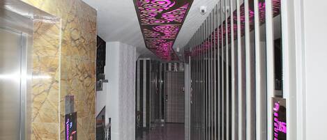 Un pasillo con pared de mármol, techo con paneles decorativos rosados y una puerta de cristal a la izquierda.