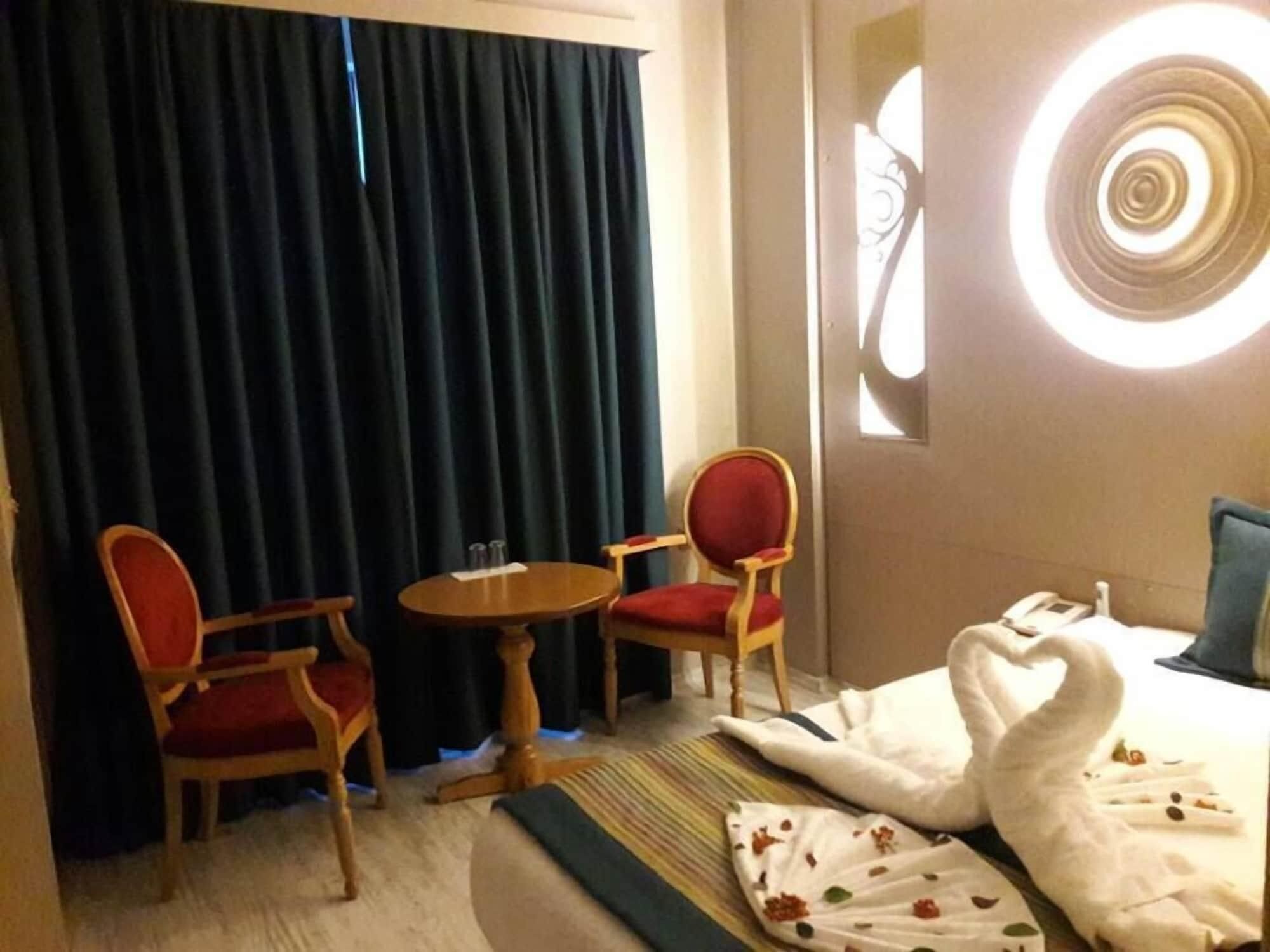 A hotel room with a bed, two red chairs, a small round table, and a window with a circular light fixture.