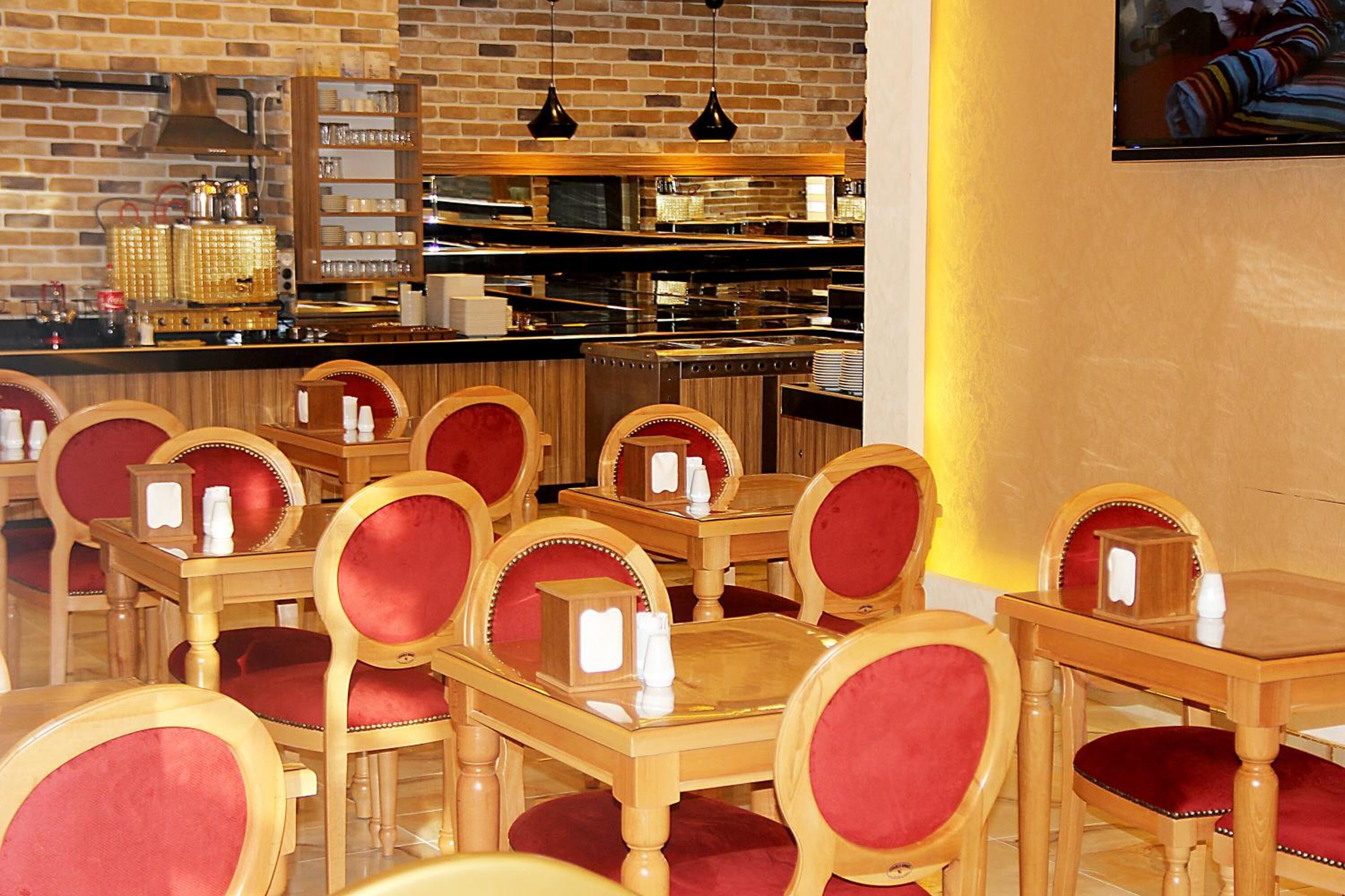 Ein Restaurantinneres mit Holztischen und rot gepolsterten Stühlen, einer Backsteinwand und einer Küche im Hintergrund.