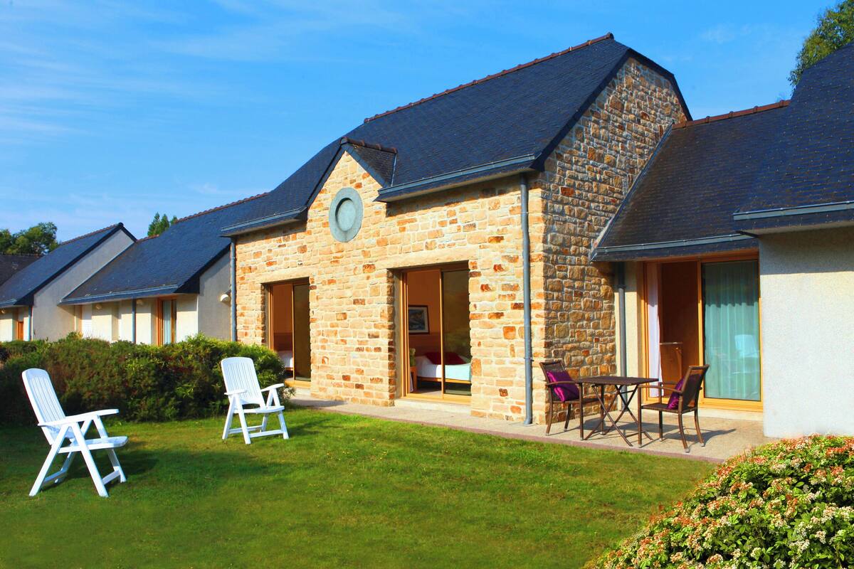A stone building with a sloped roof, a circular window, and a door leading to a room. There are two white chairs and a table outside.
