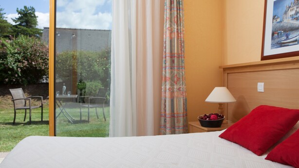 A hotel room with a large bed, a nightstand with a lamp, a framed picture on the wall, and a view of a garden and outdoor seating area. - Azureva Tregunc (Tregunc)