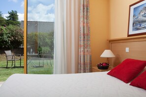 A hotel room with a large bed, a nightstand with a lamp, a framed picture on the wall, and a view of a garden and outdoor seating area.