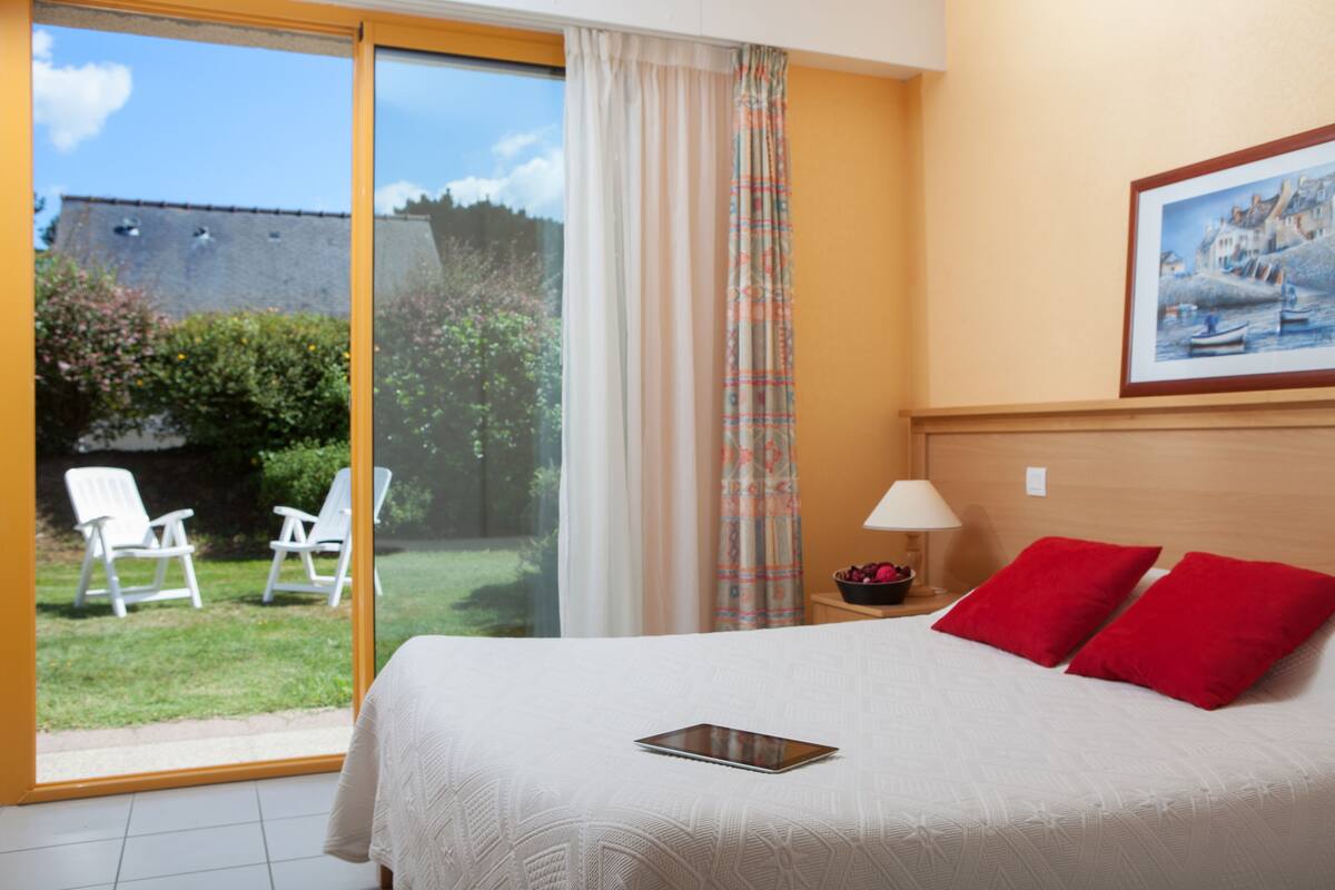 A hotel room with a bed, a tablet on the bed, a nightstand with a lamp, a painting on the wall, and a view of the outside.