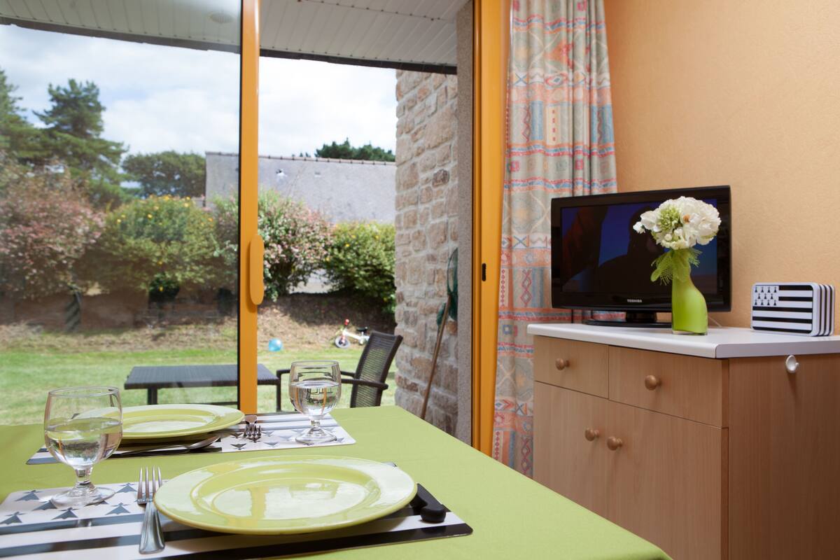 A dining table set with plates, glasses, and cutlery, a television on a cabinet, and a vase with flowers.