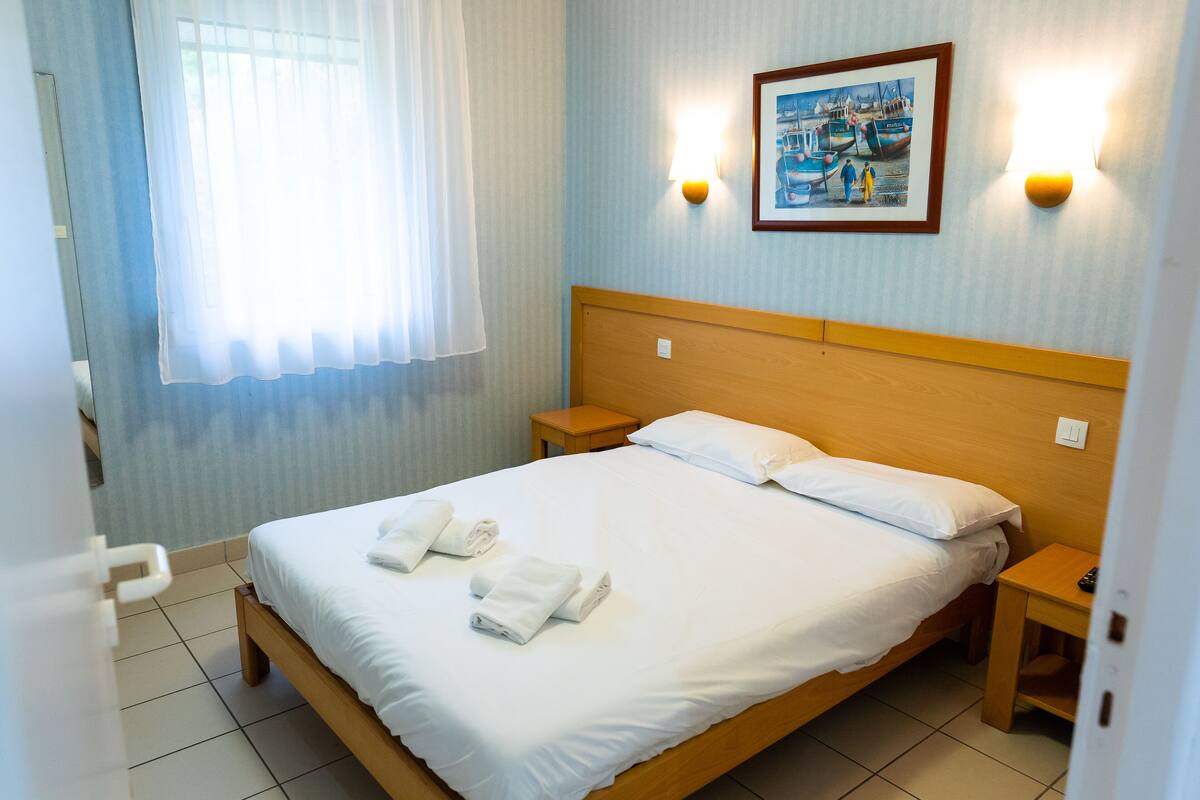 A hotel room with a bed, two bedside tables, a painting on the wall, and a window with curtains.