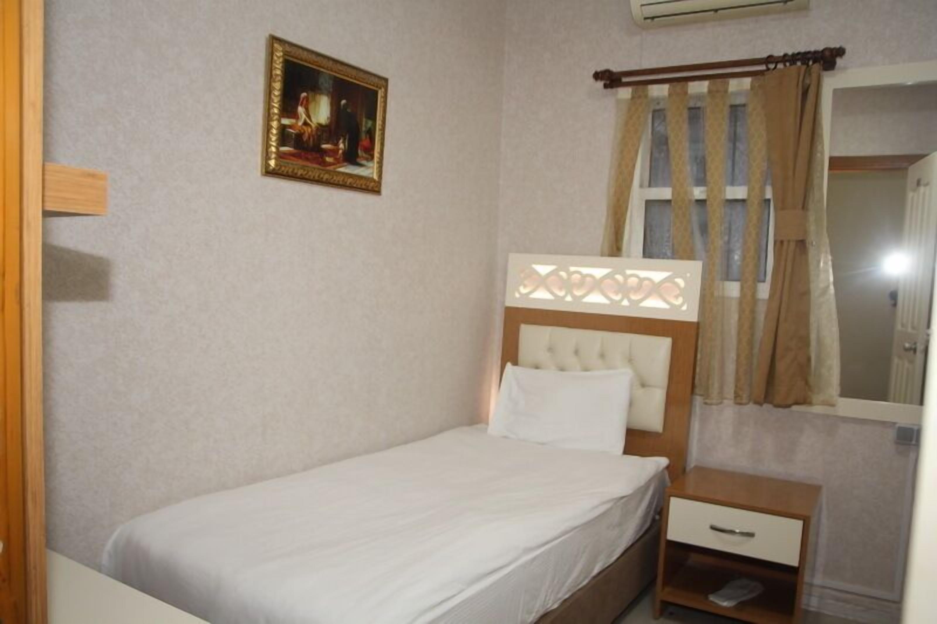 A single bed with a headboard, a bedside table, a small drawer, a framed picture on the wall, and a window with curtains.