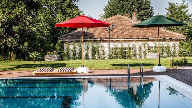 A swimming pool with two umbrellas, a white building with a tiled roof, and lush greenery in the background. - My Green Otel (Kartepe)