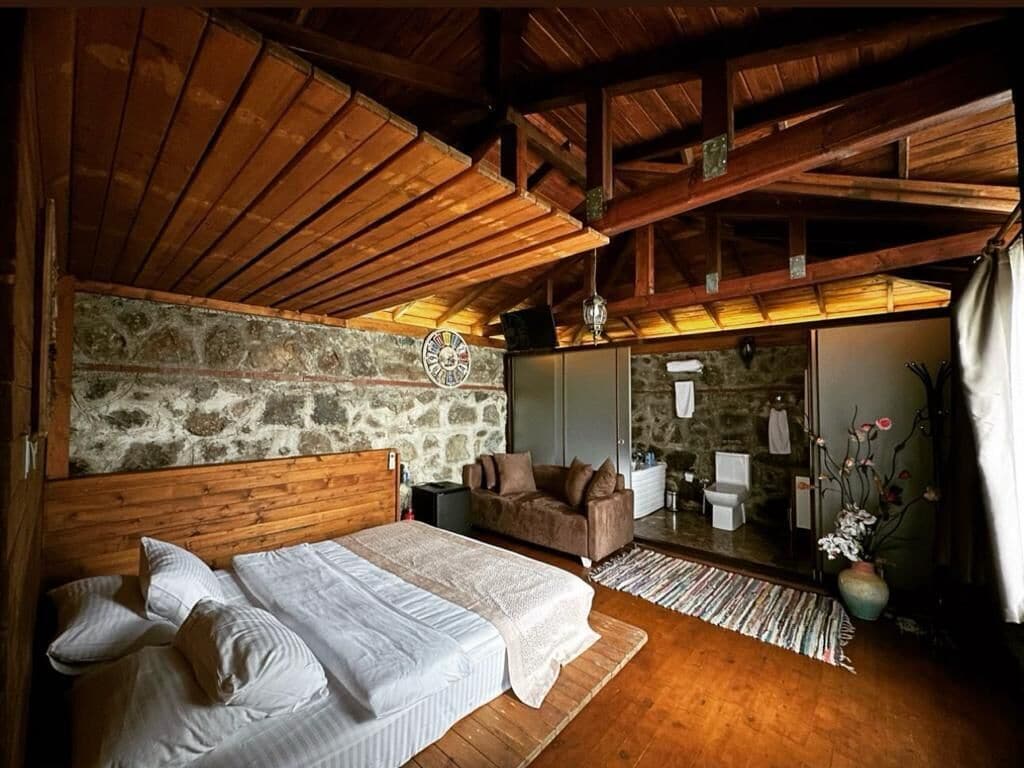 Un dormitorio acogedor con techo de madera, pared de piedra, cama con ropa blanca, sofá marrón y una pequeña mesa.