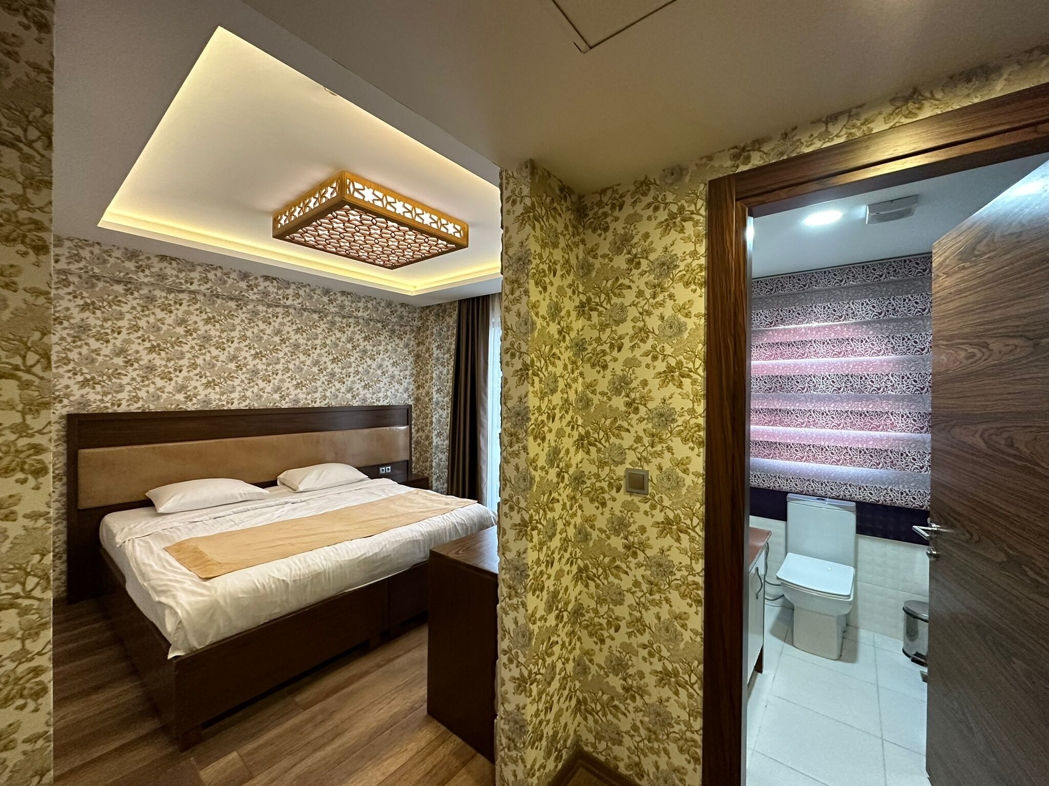 A bedroom with a bed, wooden headboard, floral wallpaper, and a ceiling light fixture.