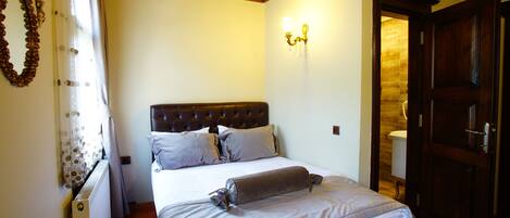 A bedroom with a wooden headboard, a bed with a rolled-up blanket, a wall-mounted light fixture, and a door leading to a bathroom.