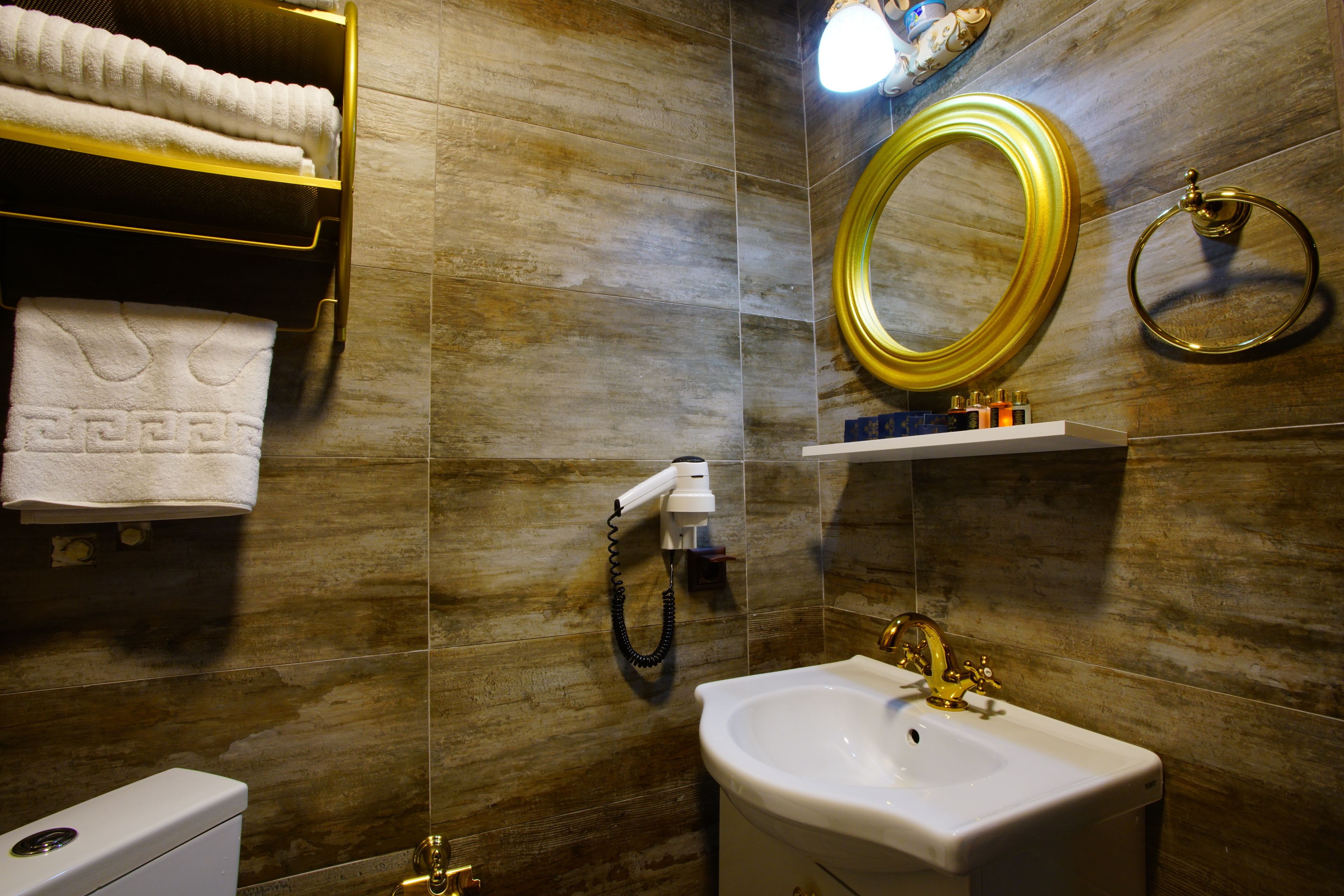 A bathroom with a white sink, gold fixtures, a towel rack with white towels, a mirror, and a towel dispenser.