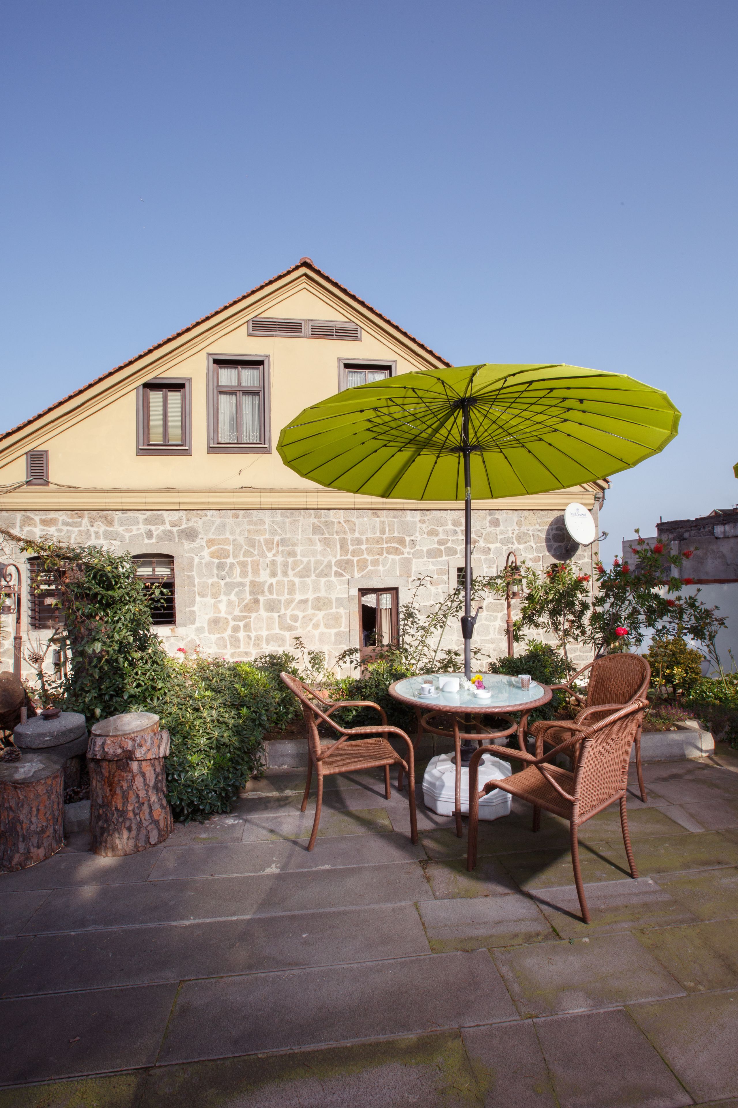 Eine Terrassengruppe im Freien mit Steinböden, einem runden Tisch, Korbstühlen und einem grünen Sonnenschirm.