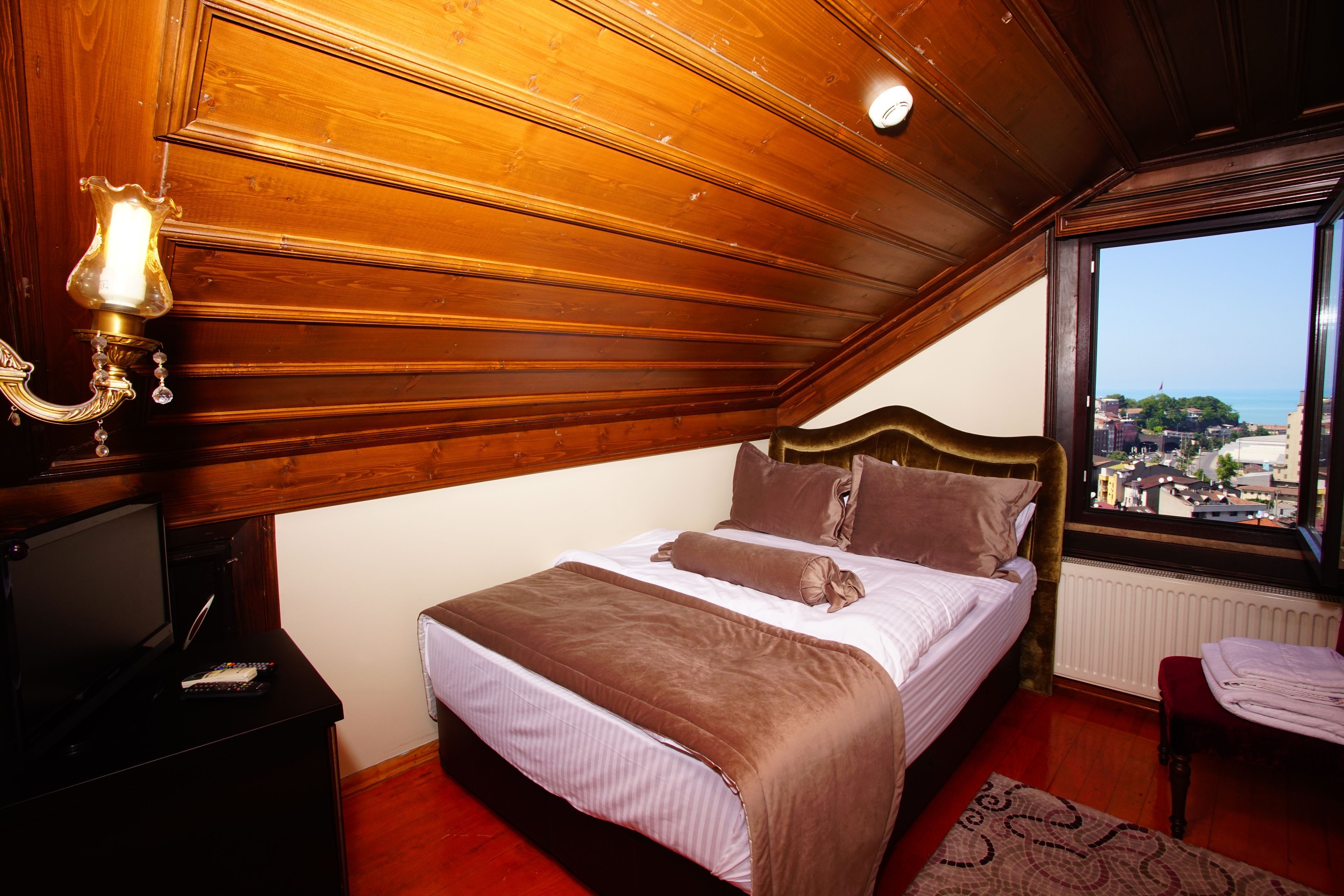 A bedroom with a wooden ceiling, a bed with brown bedding, a television on a stand, and a window with a city view.