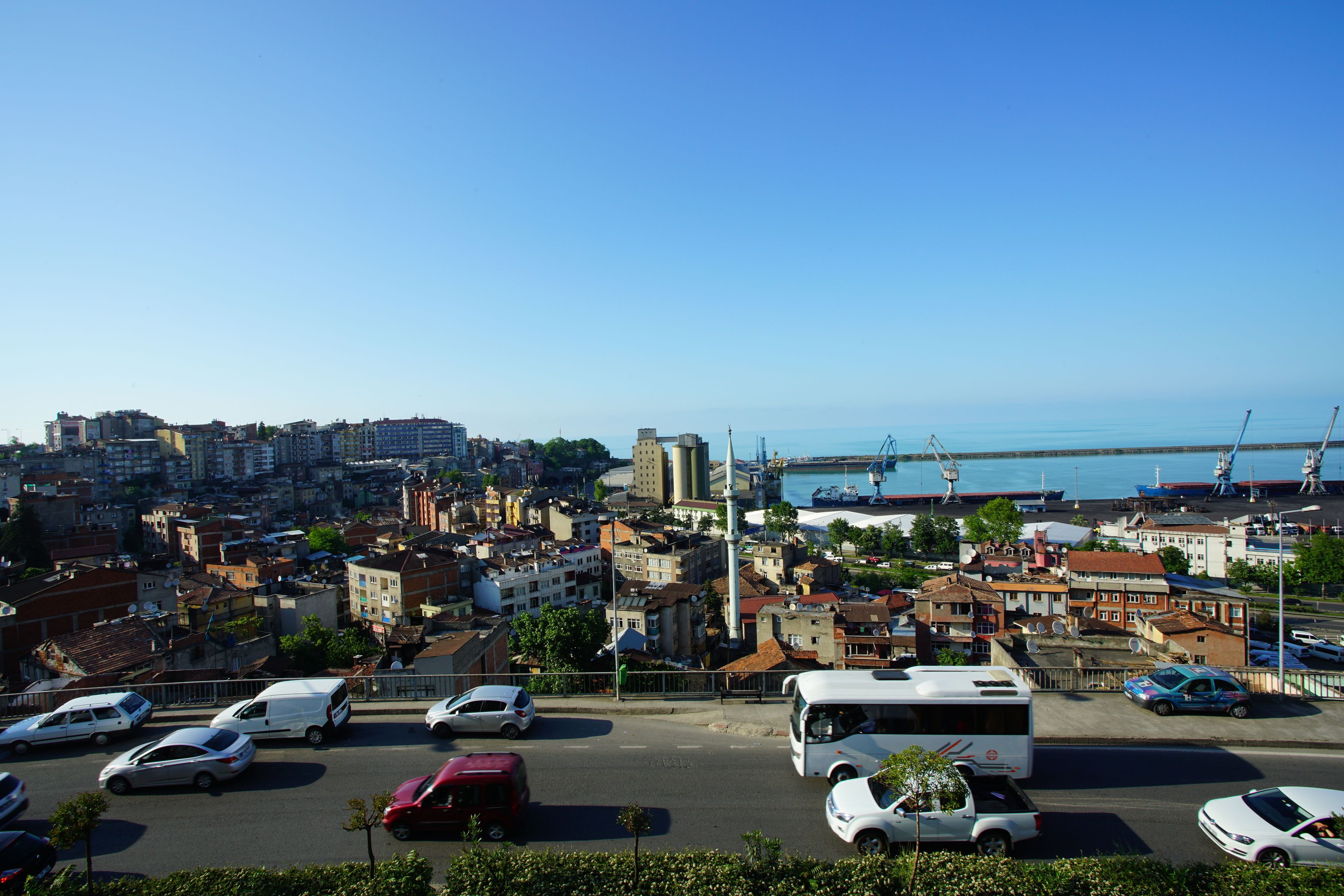 A cityscape with a view of the sea, featuring a mix of residential and commercial buildings, a bridge, and a parking lot with various vehicles.