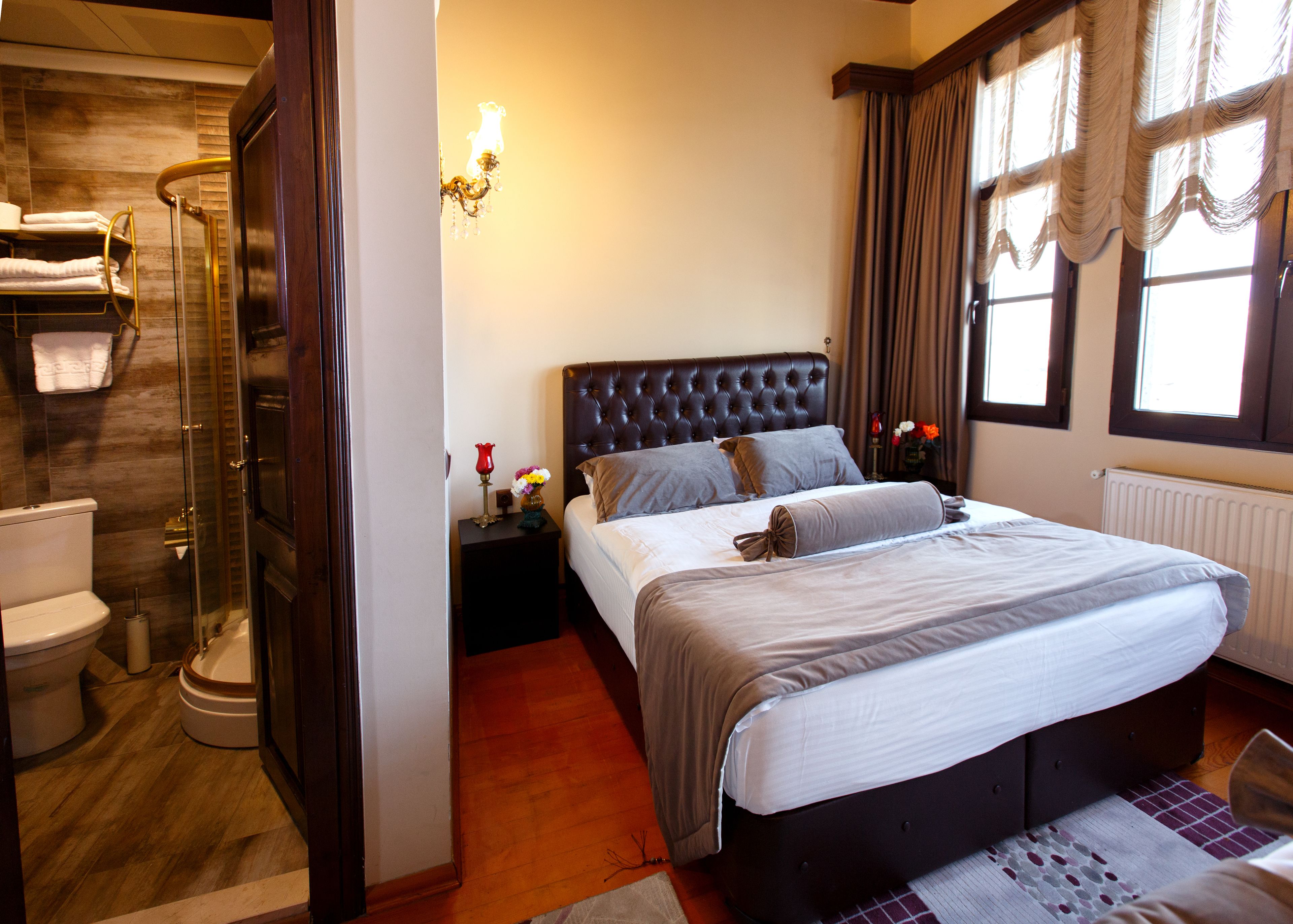 A bedroom with a large bed, a nightstand with a lamp, a bathroom with a shower, and a window with curtains.
