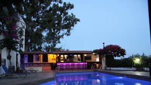Une piscine extérieure avec des chaises longues, un espace bar éclairé de lumières violettes et un arbre aux fleurs roses.