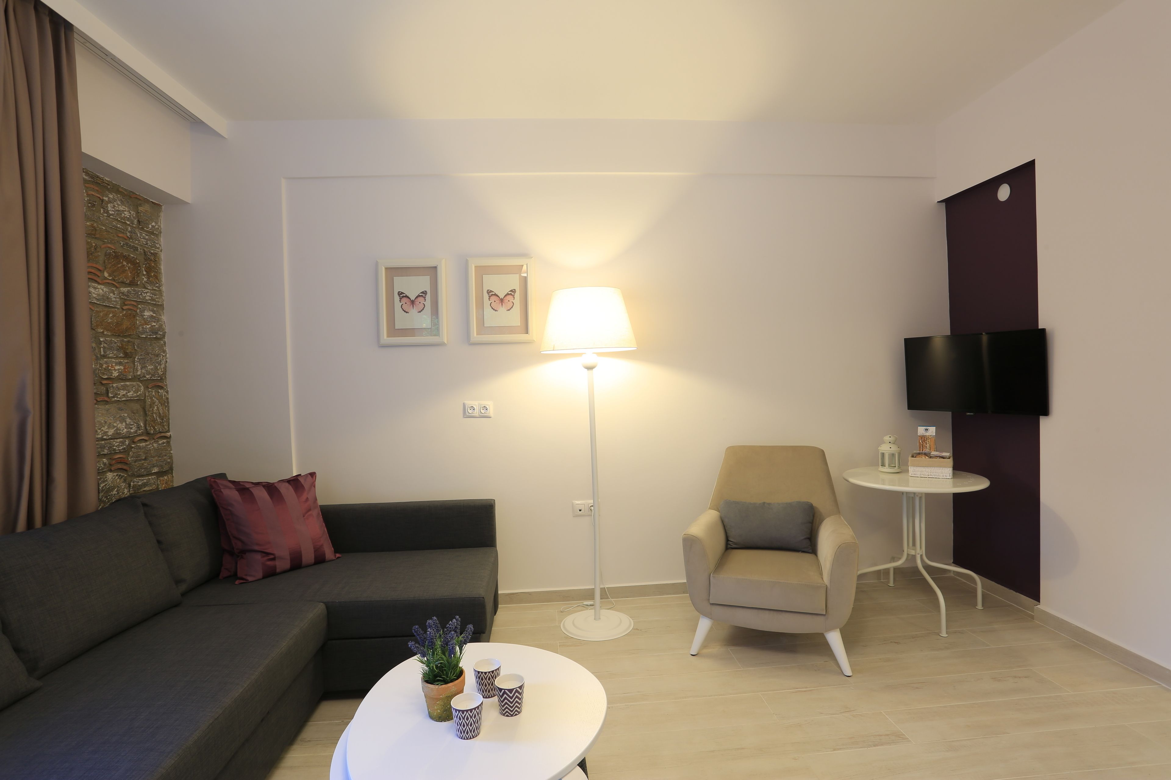 A modern living room with a grey sofa, a beige armchair, a small round table, a floor lamp, and a flat-screen TV mounted on the wall.