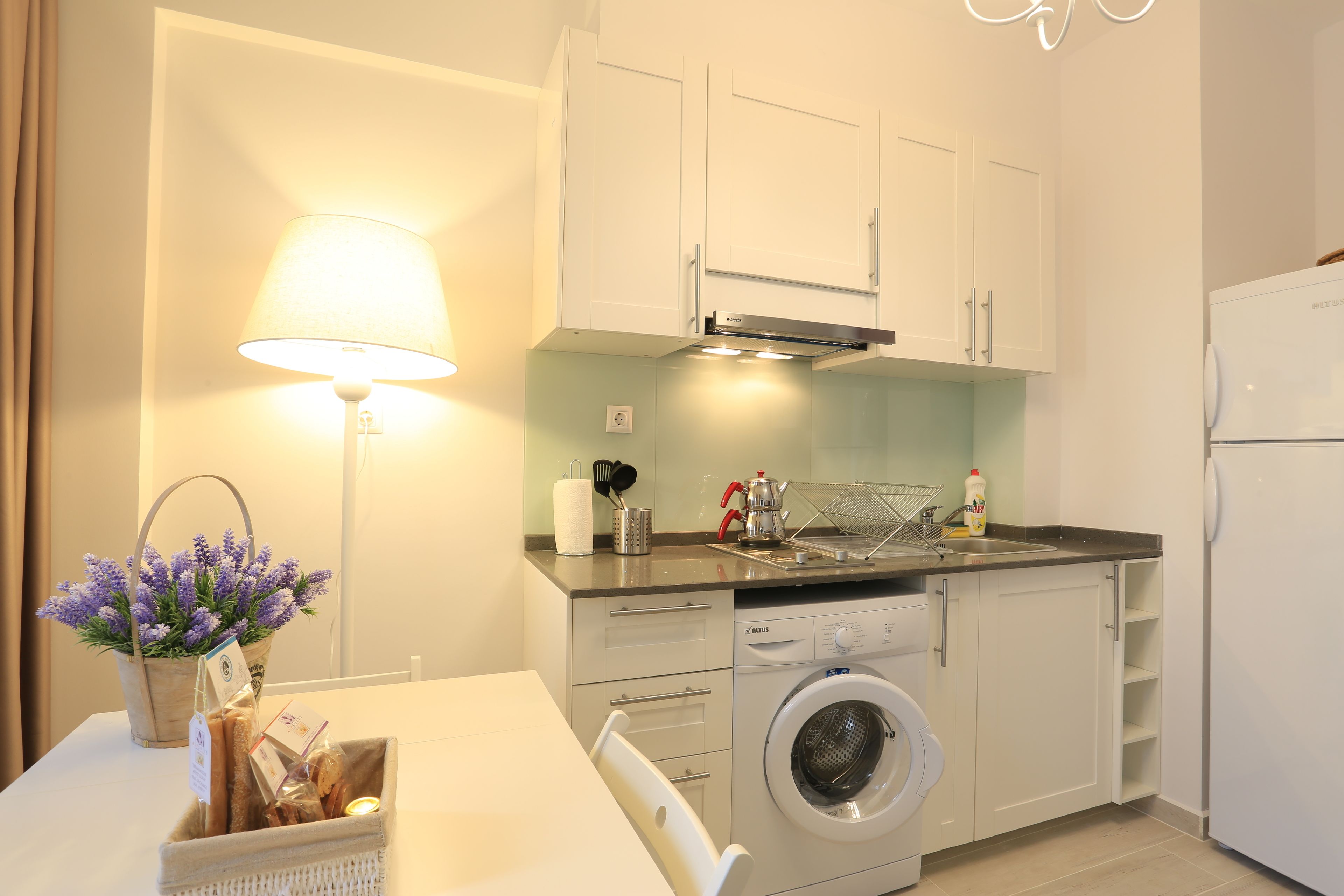 A modern kitchen with white cabinets, a washing machine, a sink, and a table with a basket of bread and a lamp.