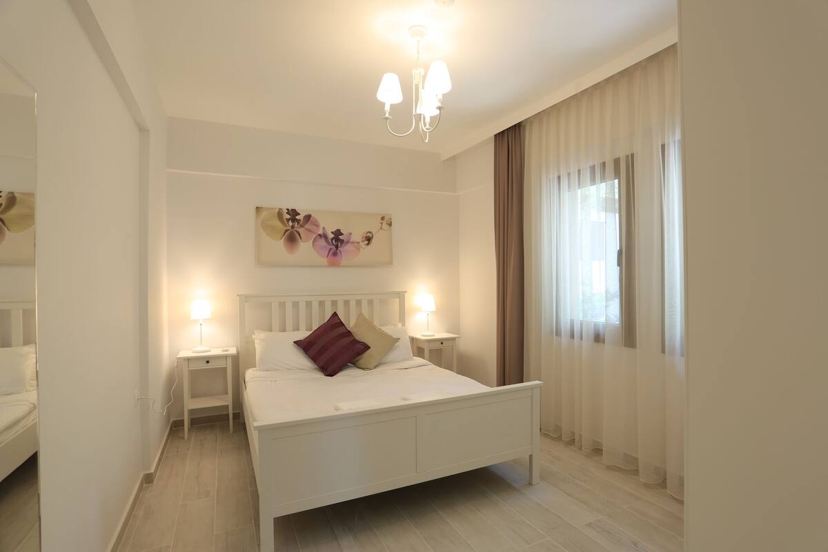 Une chambre à coucher avec un lit blanc, deux tables de chevet avec des lampes, un tableau au-dessus du lit et une fenêtre avec des rideaux.