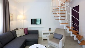 A modern living room with a grey sofa, a beige armchair, a white coffee table, a flat-screen TV, a white desk, and a wooden spiral staircase. - Alp Suites Lavanta (Ula)