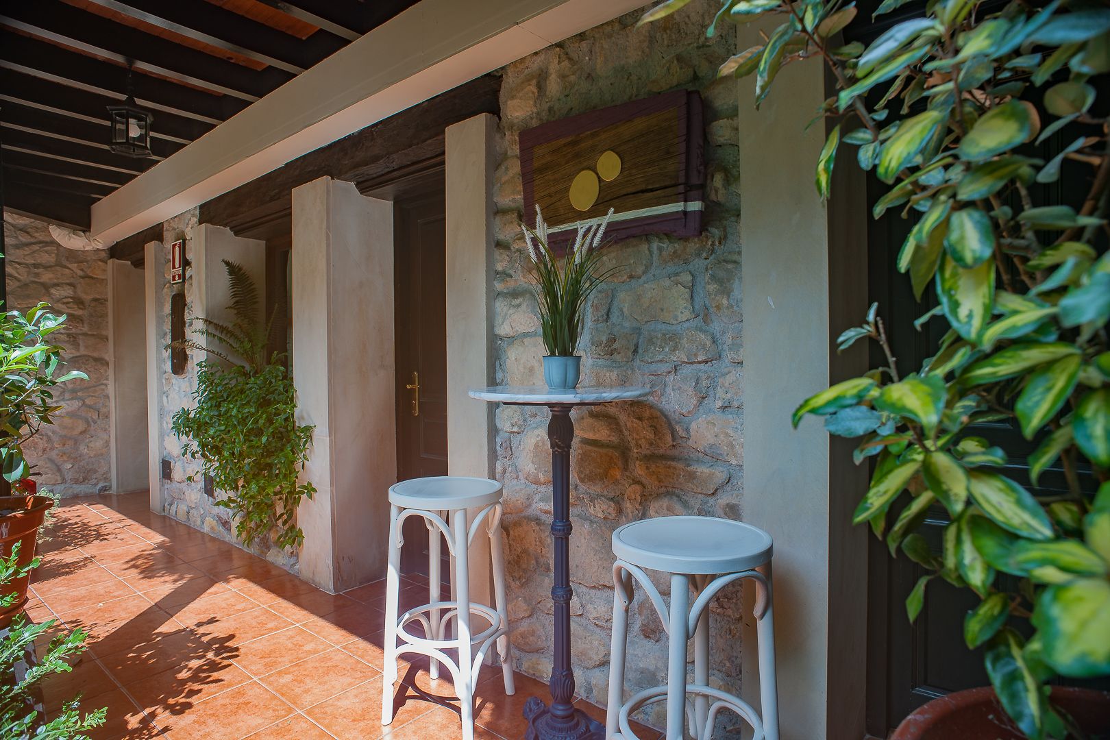 A stone wall with a wooden shelf holding a potted plant and a framed picture. There are two white stools and a small table with a potted plant.