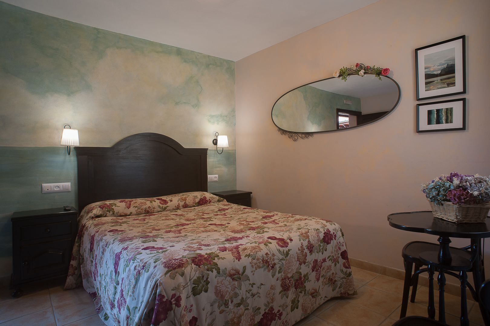 A bedroom with a floral bedspread, a round mirror, and two wall-mounted lamps.