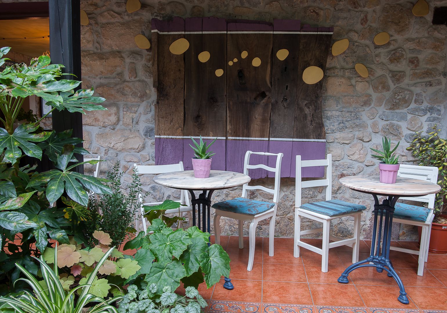 Un patio al aire libre con paredes de piedra, sillas y mesas blancas con plantas en macetas.