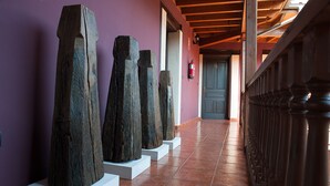 Un pasillo con esculturas de madera sobre pedestales, un extintor en la pared y una puerta al fondo.