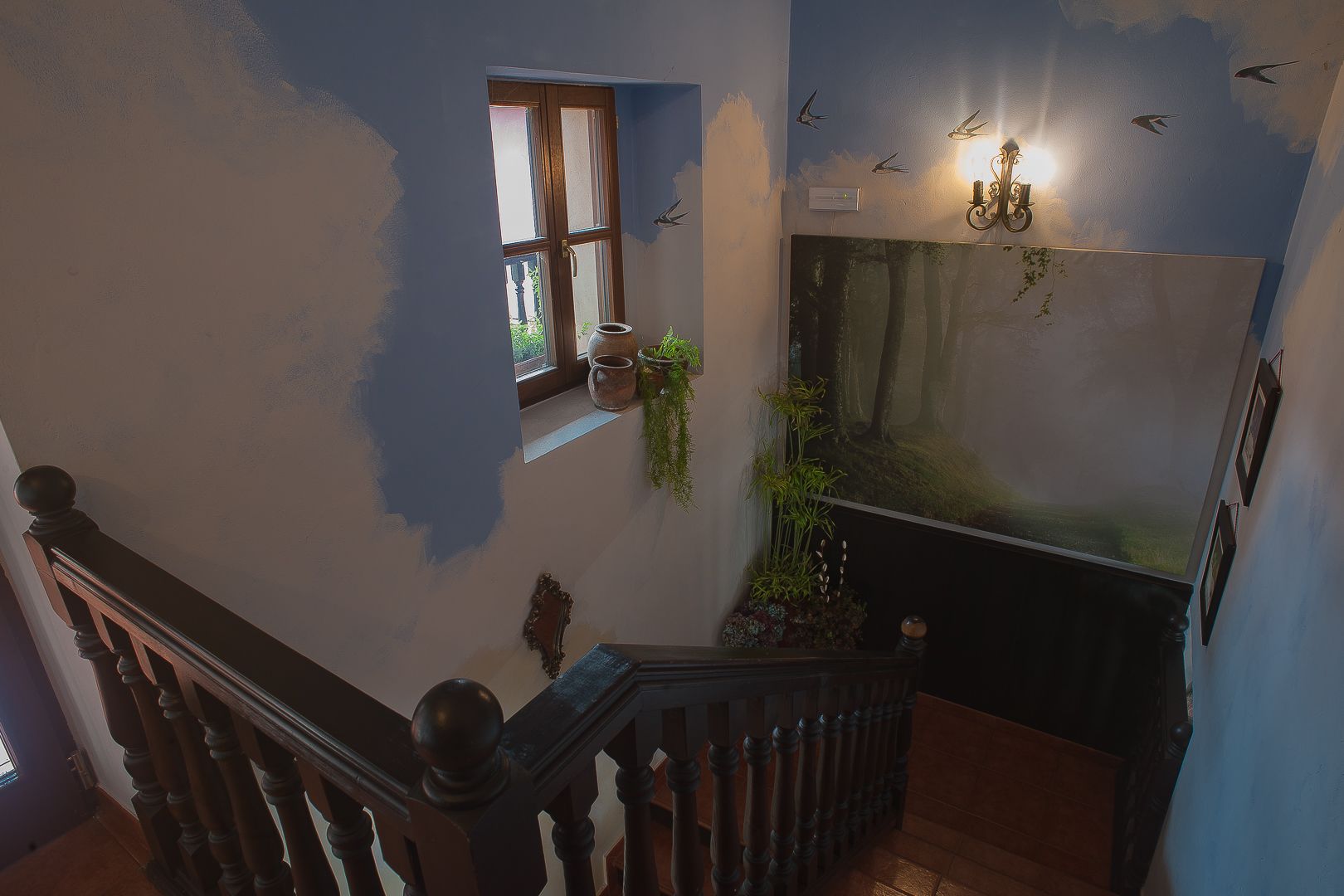 Una escalera con barandilla de madera, un ventanal con plantas y una pared con un mural y luces empotradas.