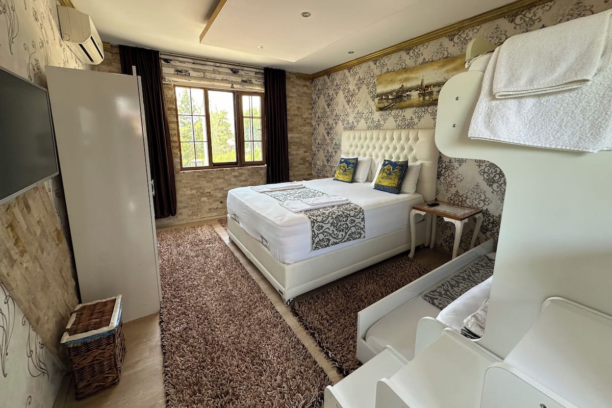 A bedroom with a large bed, a bedside table, a wardrobe, a window with curtains, and a wall with a decorative pattern.