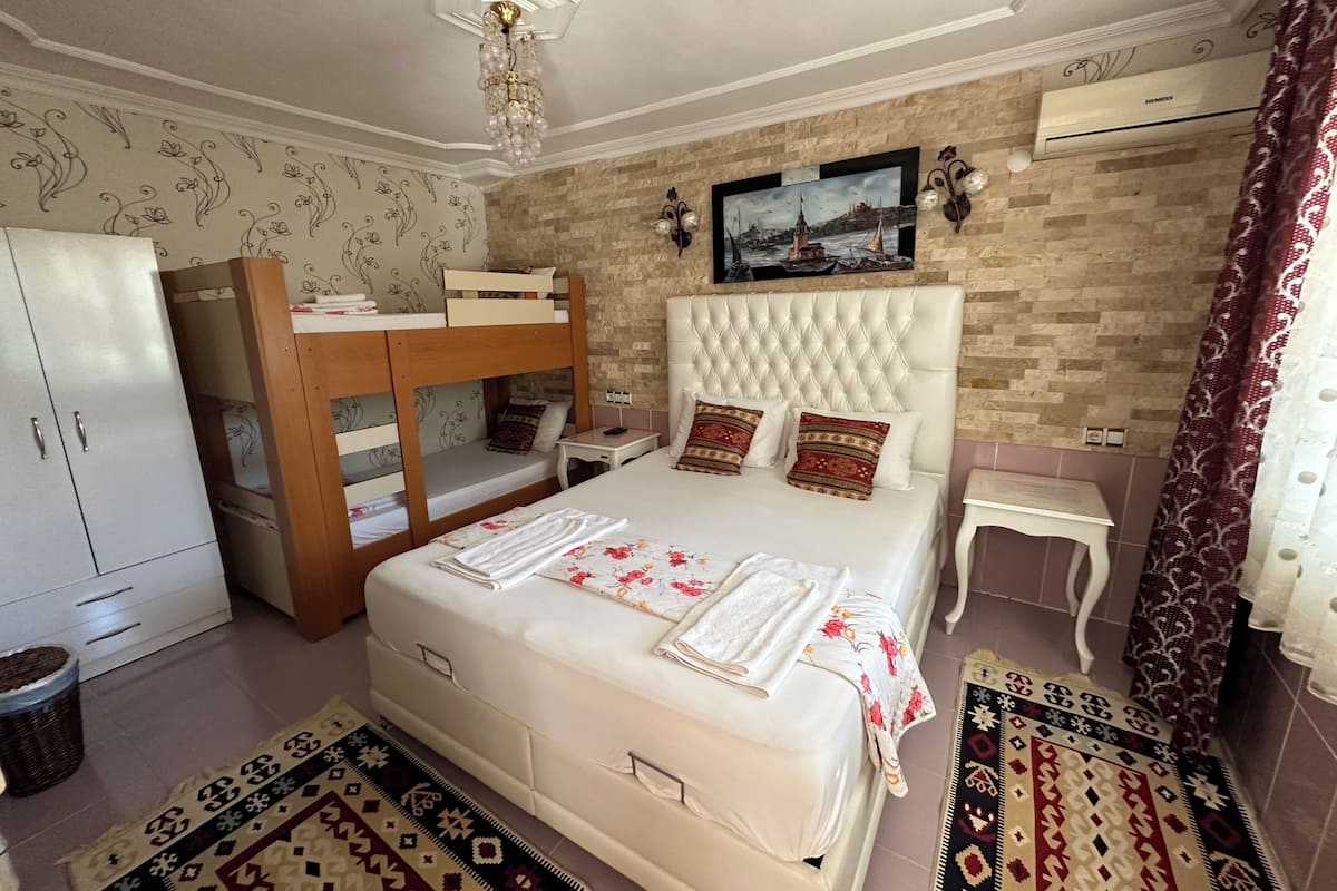 A bedroom with a bunk bed, a large bed with pillows, a nightstand, a wall-mounted air conditioner, and patterned rugs.