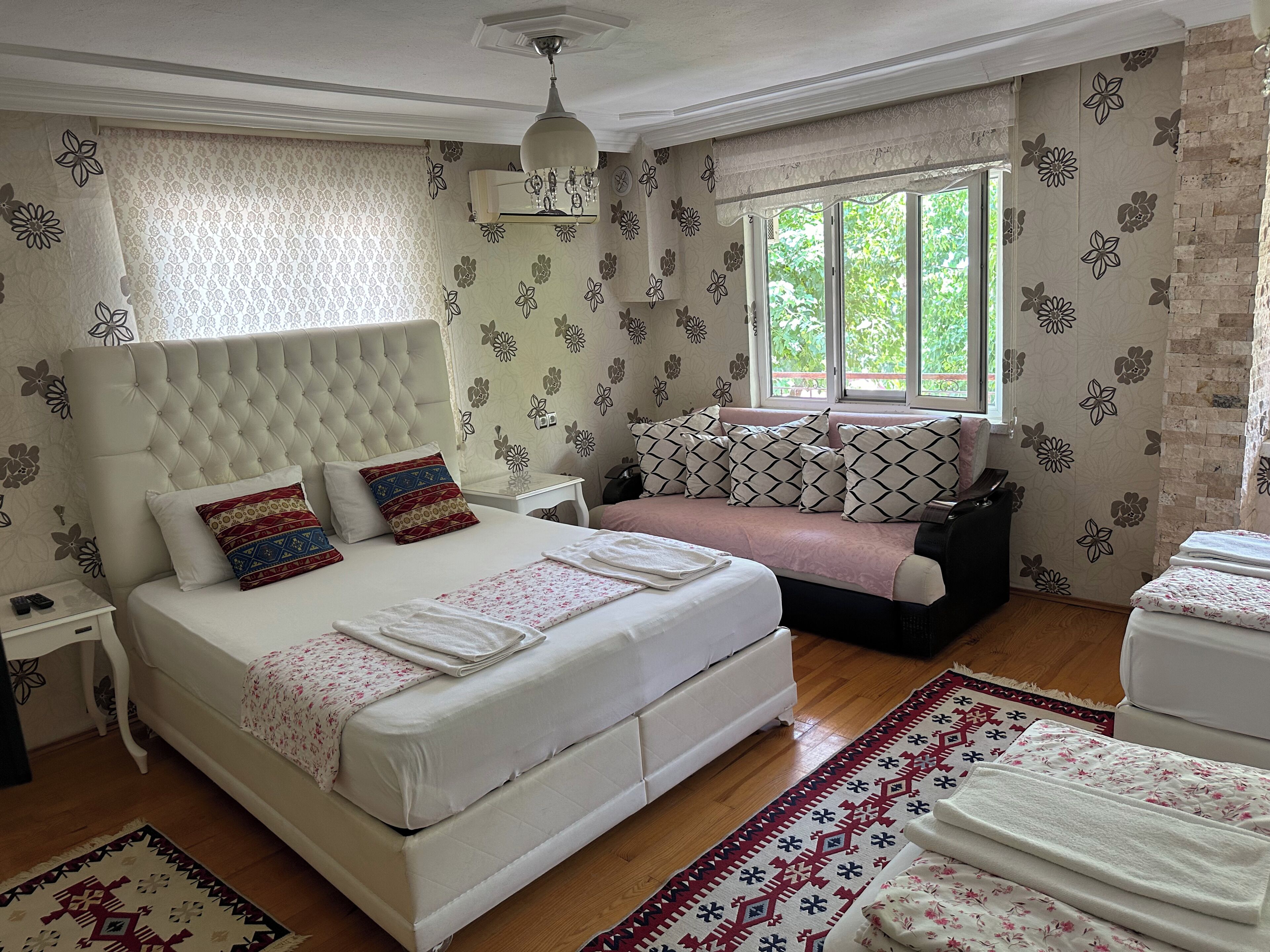 A bedroom with a large bed, a sofa, a small table, and a window with a view of greenery.
