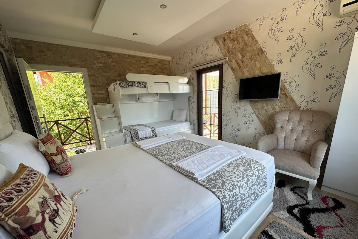 A bedroom with a large bed, a chair, a TV, and a balcony with a view of greenery.