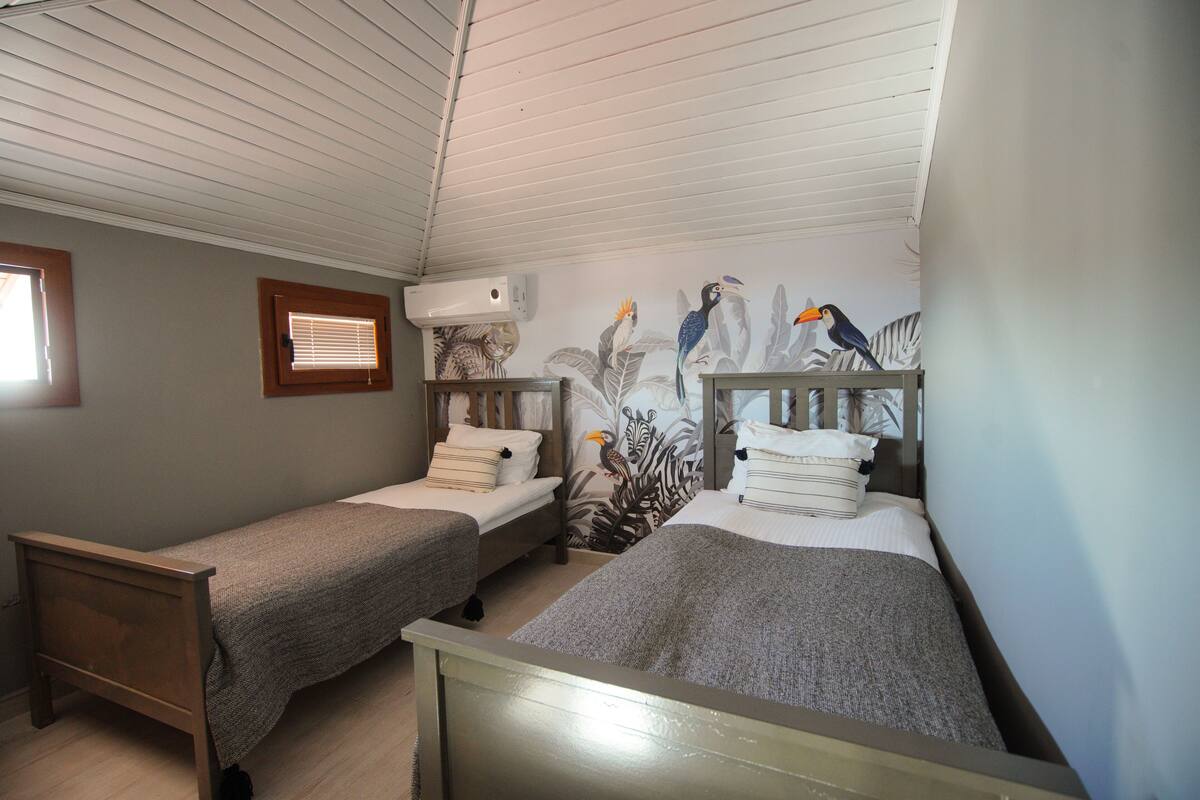 Una habitación con dos camas individuales, techo de madera y un mural en la pared con pájaros y follaje.