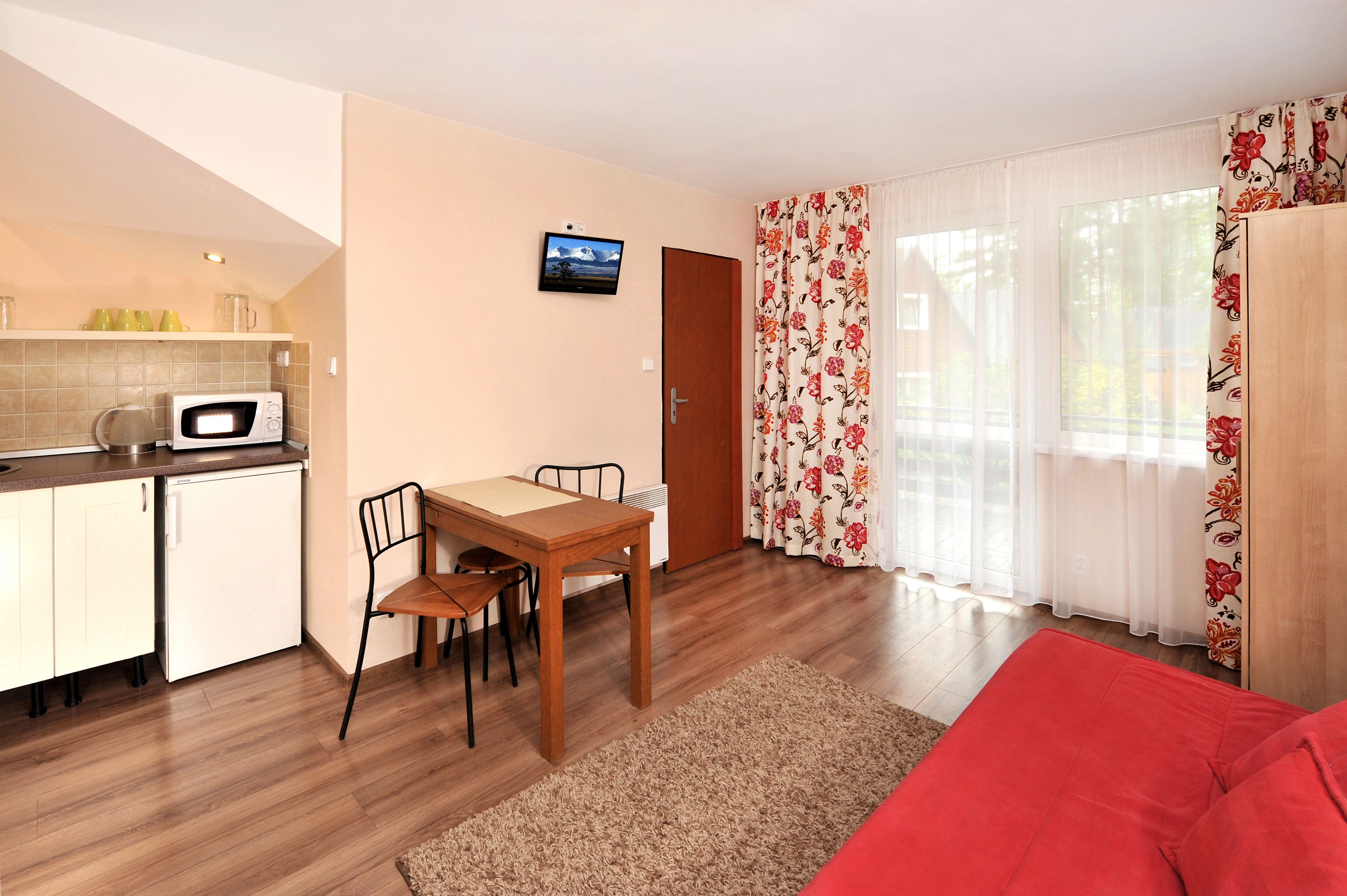 Een ruimtelijke woonruimte met een kitchenette, een eethoek met tafel en stoelen, een rode bank en een aan de muur bevestigde televisie.