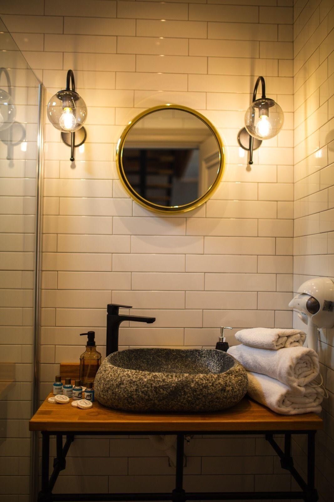 Un baño con un espejo redondo, dos luces empotradas, un grifo negro, un lavabo de piedra y un perchero con toallas.