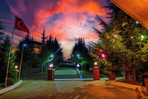 A paved entrance with steps leading up to a building, surrounded by trees and a flag. - Akcatepe Dag Evleri (Akçaabat)