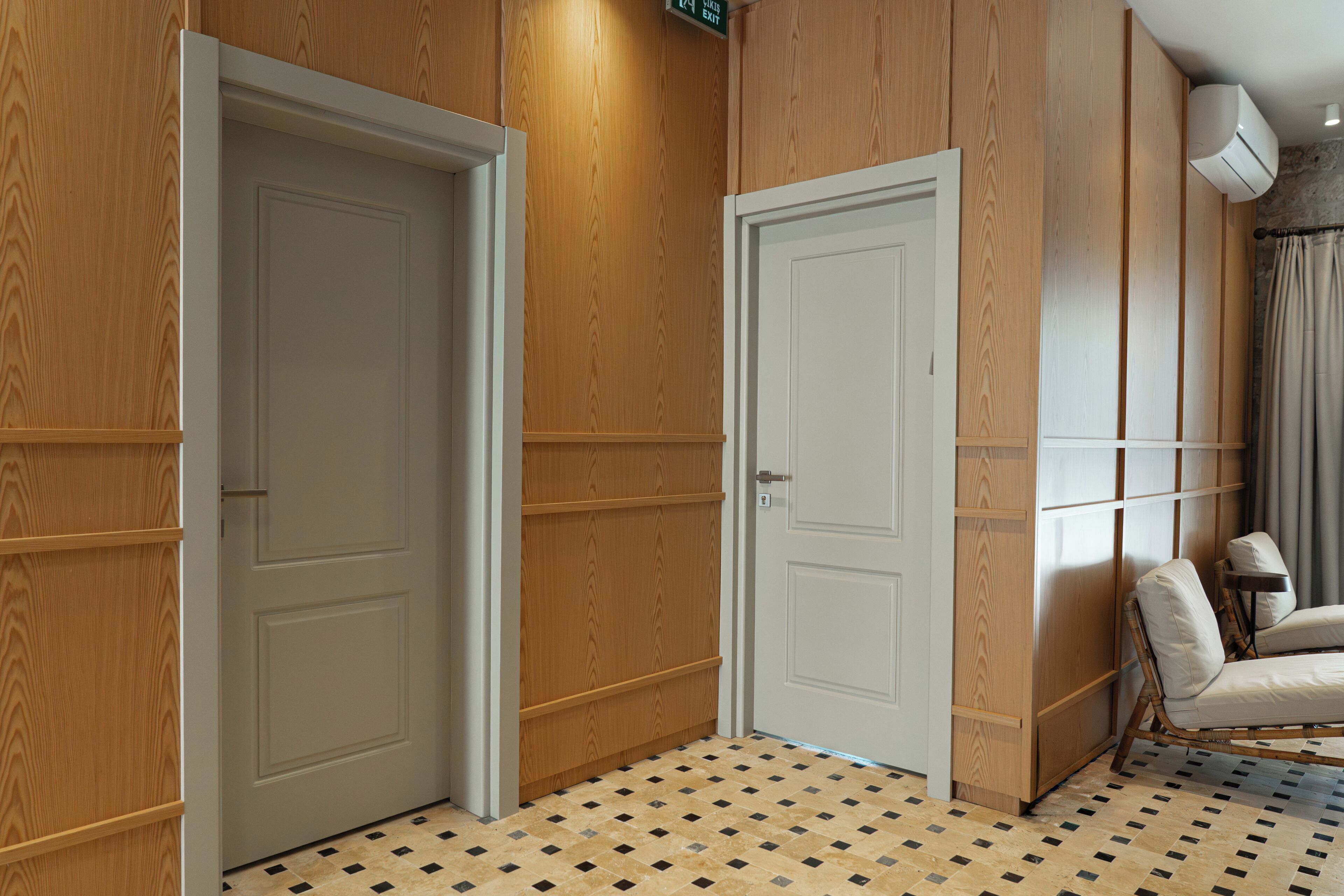 Habitación de hotel con dos puertas, suelo de baldosas, pared de paneles de madera, unidad de aire acondicionado blanca y una silla.