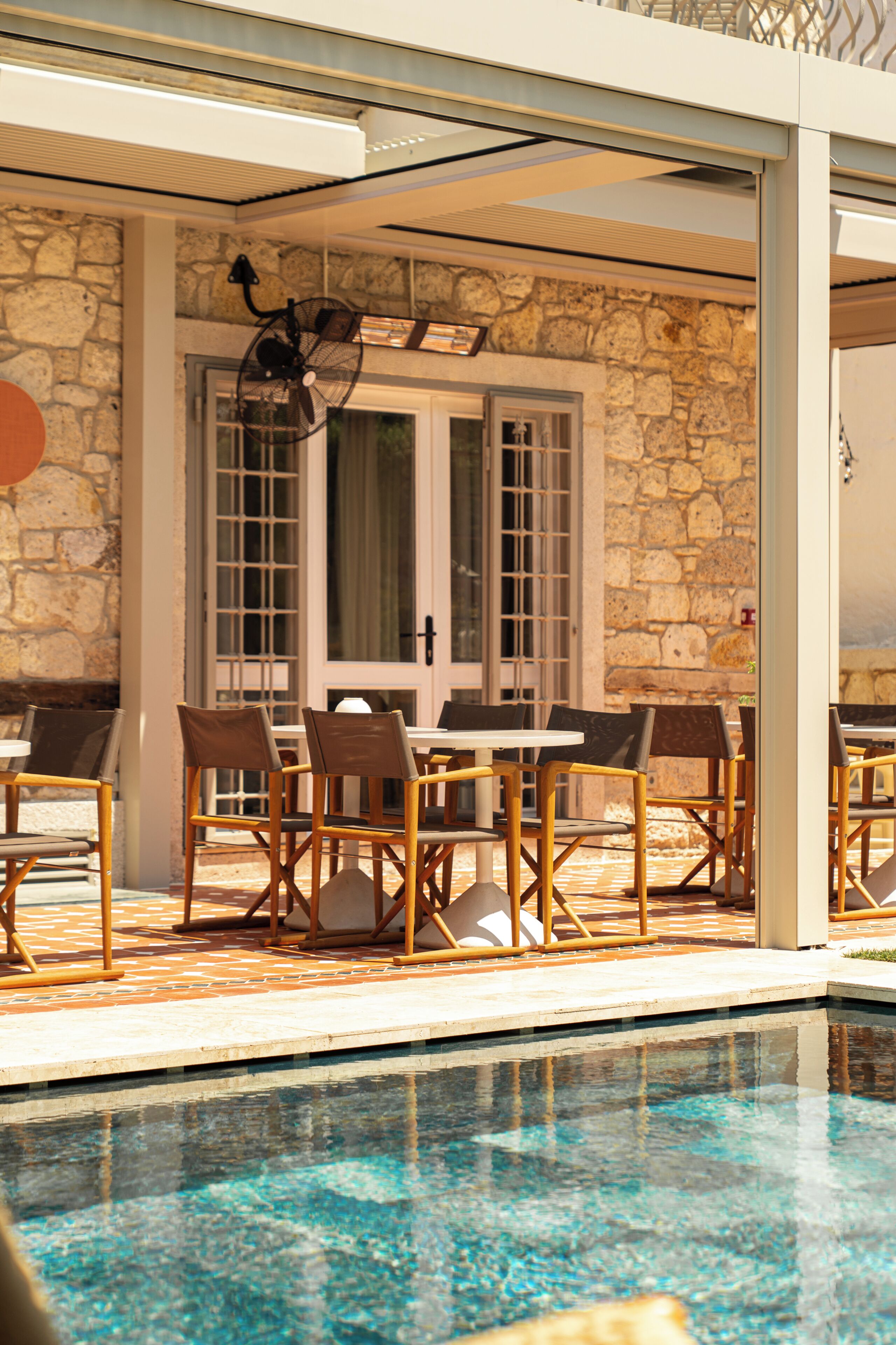 An outdoor pool area with stone walls, a glass door, and wooden chairs and tables.