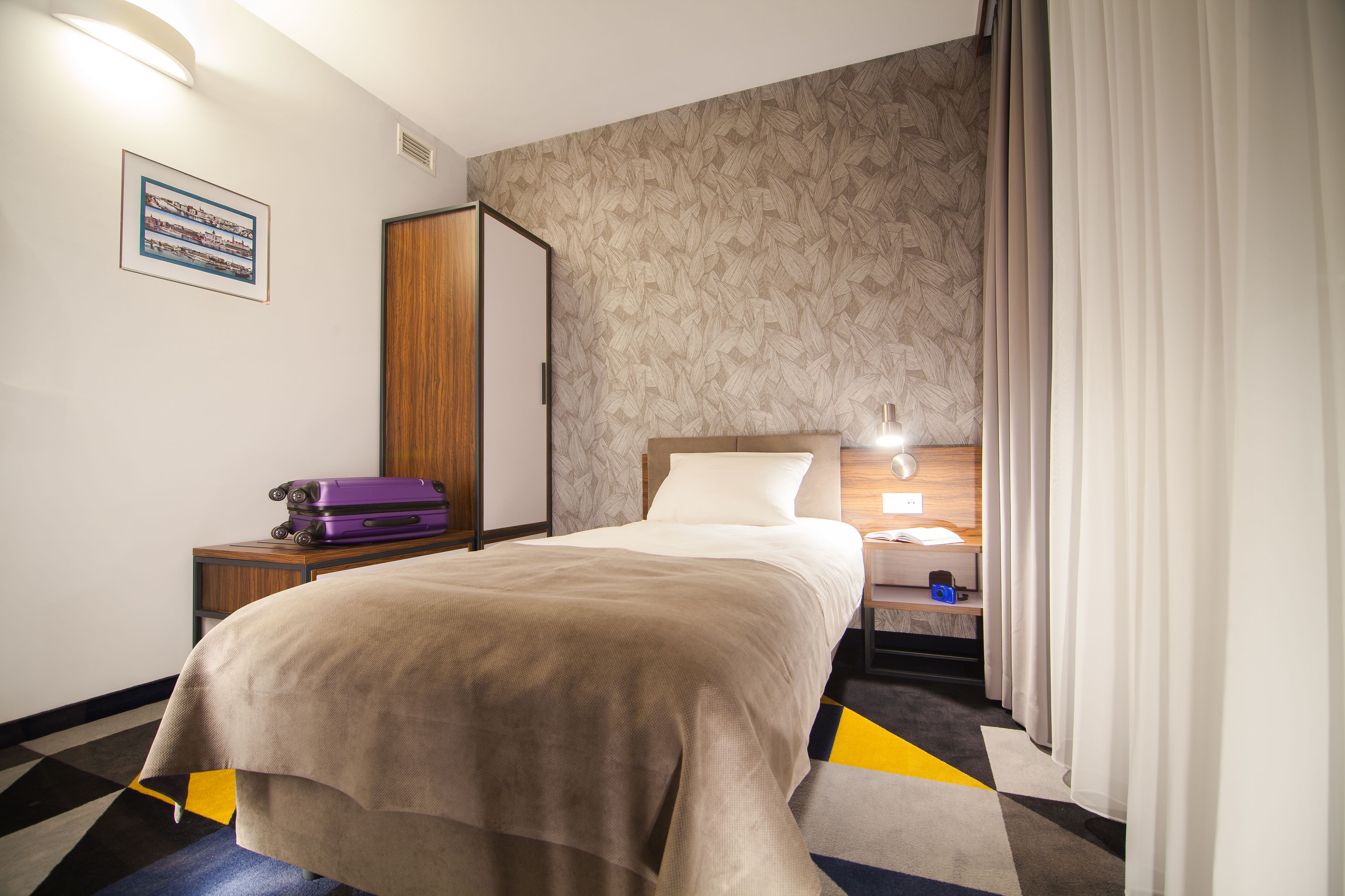 Ein Hotelzimmer mit Bett, Nachttisch, Koffer, Kleiderschrank und gemustertem Wandpapier.
