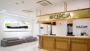 A hotel reception area with a wooden reception desk, a large window with a cityscape view, and a VULCAN hotel sign. - Vulcan Hotel (Szczecin)