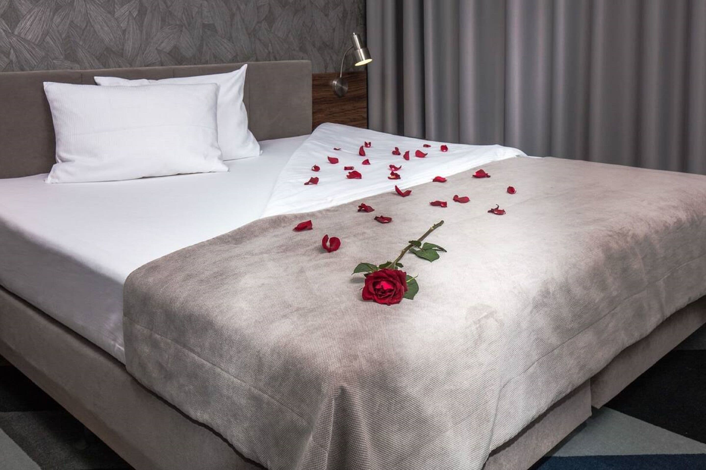 A neatly made bed with white linens, a grey throw blanket, and scattered rose petals and a single rose on the bed.