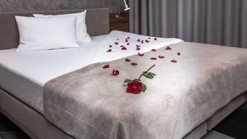 A neatly made bed with white linens, a grey throw blanket, and scattered rose petals and a single rose on the bed.